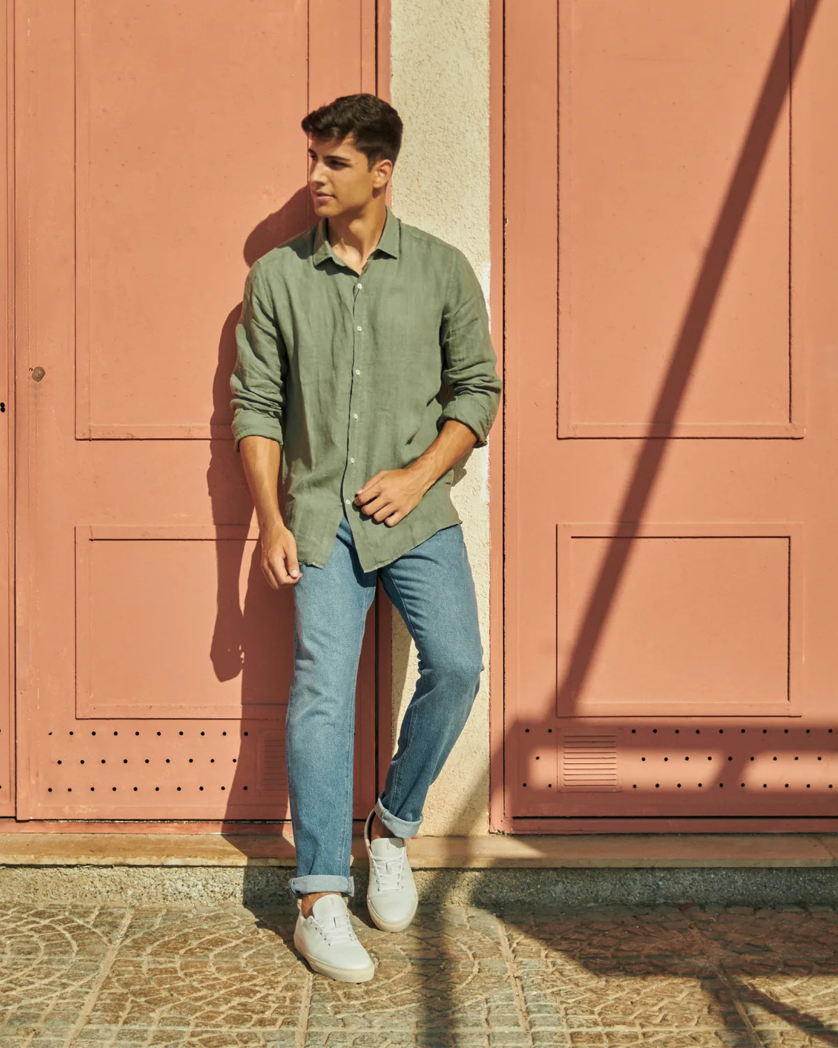 Hombre apoyado en una puerta rosa con camisa de lino verde y vaqueros llevando las Bauhaus pure white